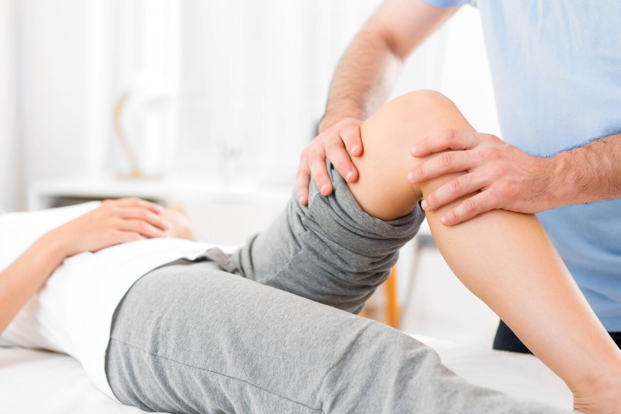 Physiotherapeut untersucht das rechte Knie einer liegenden Person zur Diagnose von Knieschmerzen – typisches Szenario bei Gonalgie, etwa nach Überlastung, Arthrose oder Verletzungen.